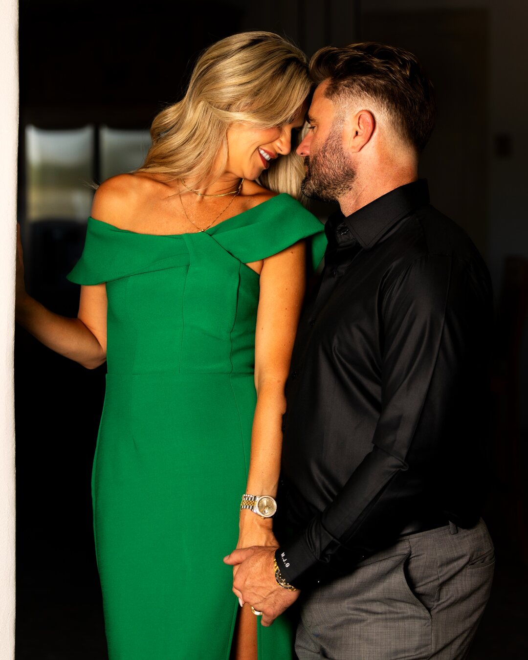 A couple stand close with their foreheads resting together and hands intertwined, she in an emerald green dress, sharing a quiet, intimate moment.
